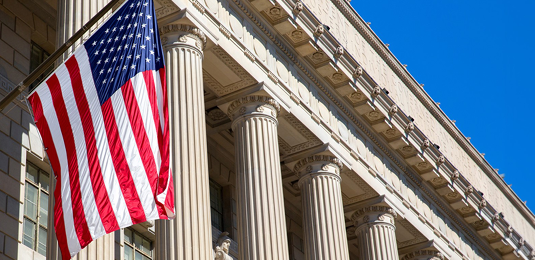 American flag in front of the U.S. Supreme Court Building with tall neoclassical columns and a clear blue sky.