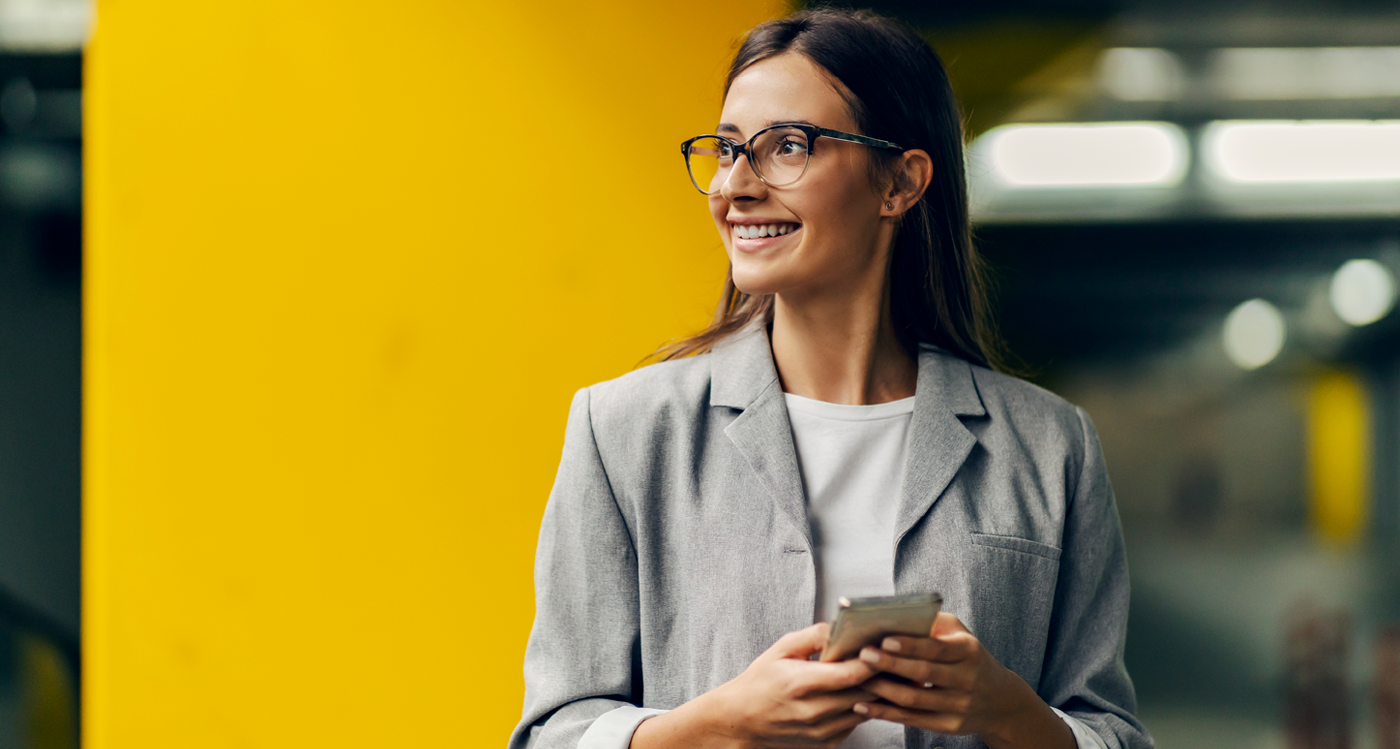 Person in a gray blazer holding a smartphone, standing in front of a yellow wall.